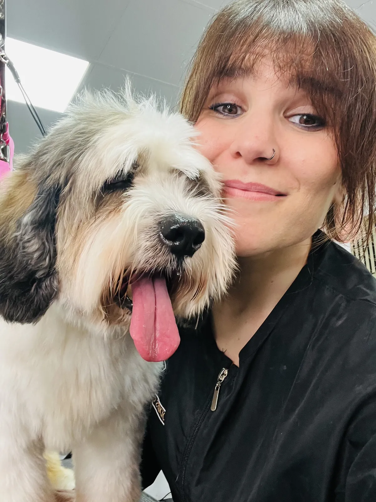 A groomer smiles next to a shaggy terrier with its tongue out
